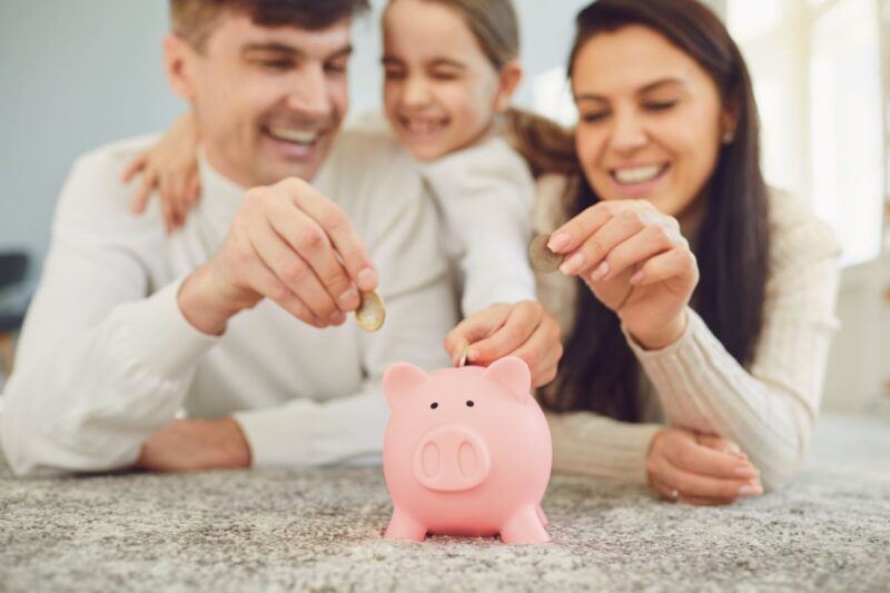 Pais com criança pequena, colocando moedas em um cofrinho de porquinho juntos, sorrindo em casa.